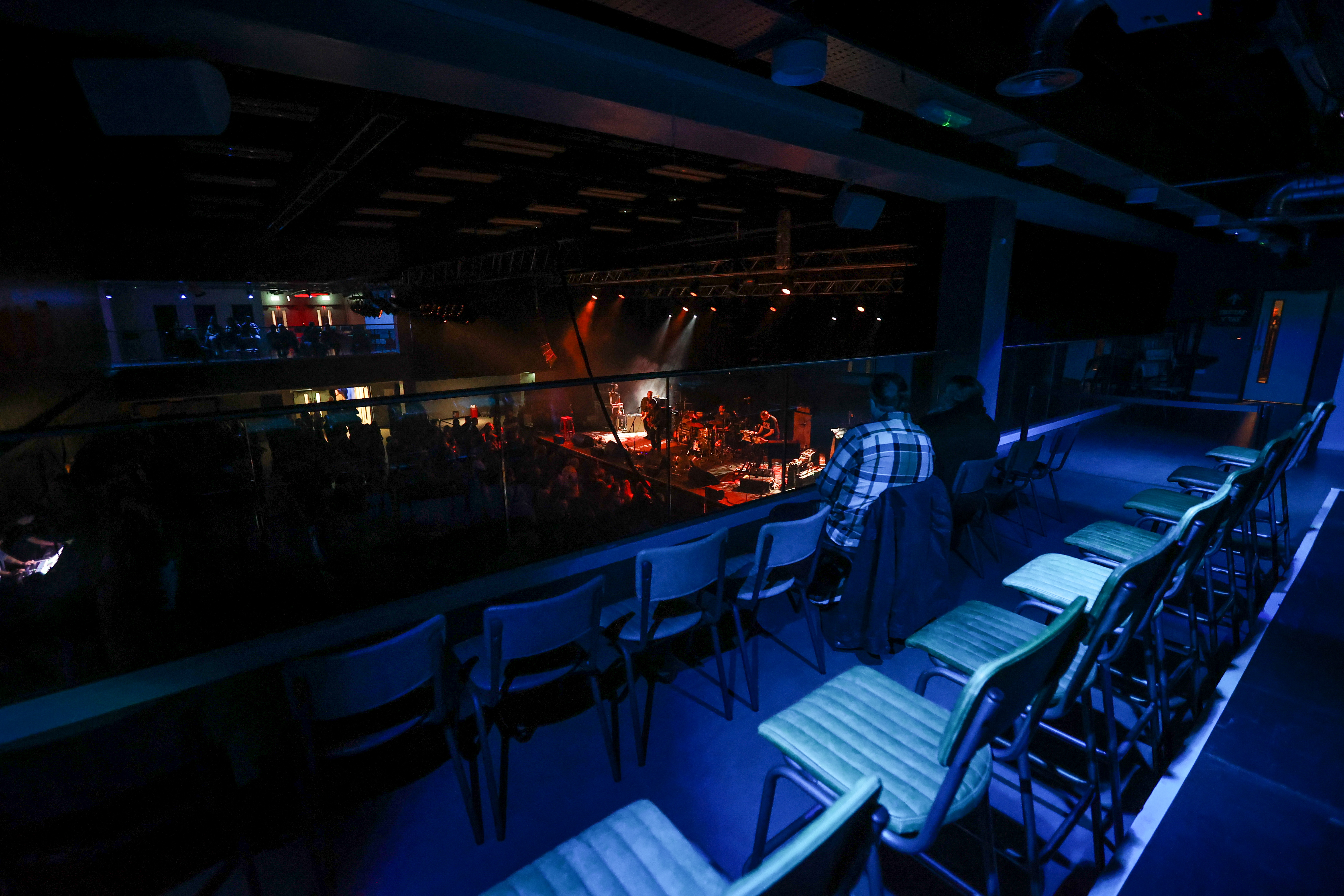 A photograph showing the accessible viewing balcony taken from a balcony overlooking a stage in a music venue. The venue is dimly lit, with the main lighting focused on the stage. The stage is brightly illuminated with red and white lights, highlighting a band performing. The audience is visible below, appearing as a crowd of silhouetted figures. The balcony features rows of chairs, arranged in a tiered formation. The balcony railing is visible in the foreground, with a person standing near it.