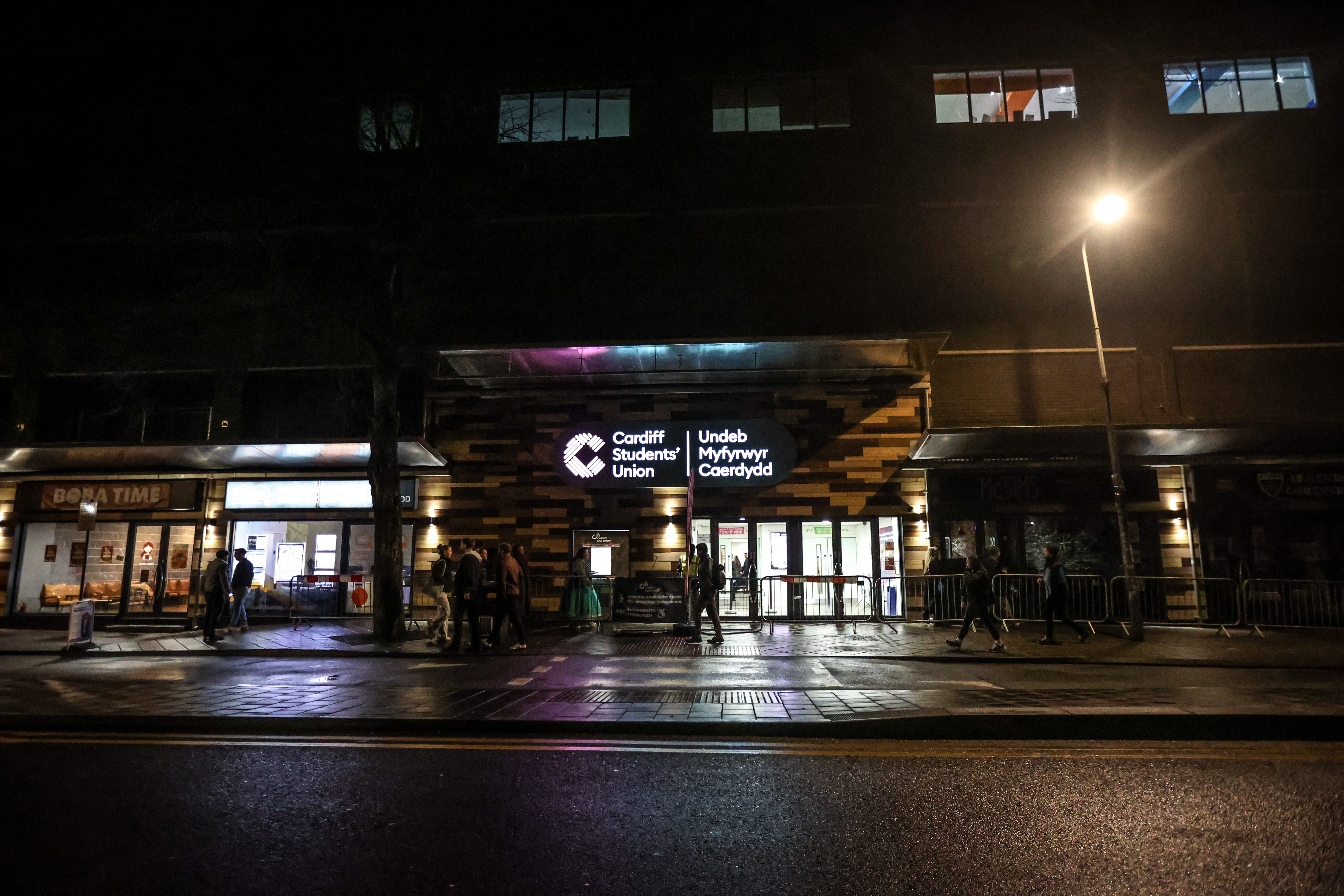 A nighttime photograph of the Cardiff Students' Union building. The building features a prominent, brightly lit entrance with glass doors and a large sign displaying 'Cardiff Students' Union'. The building facade has several illuminated windows on the upper floors. A tall streetlamp provides additional lighting.