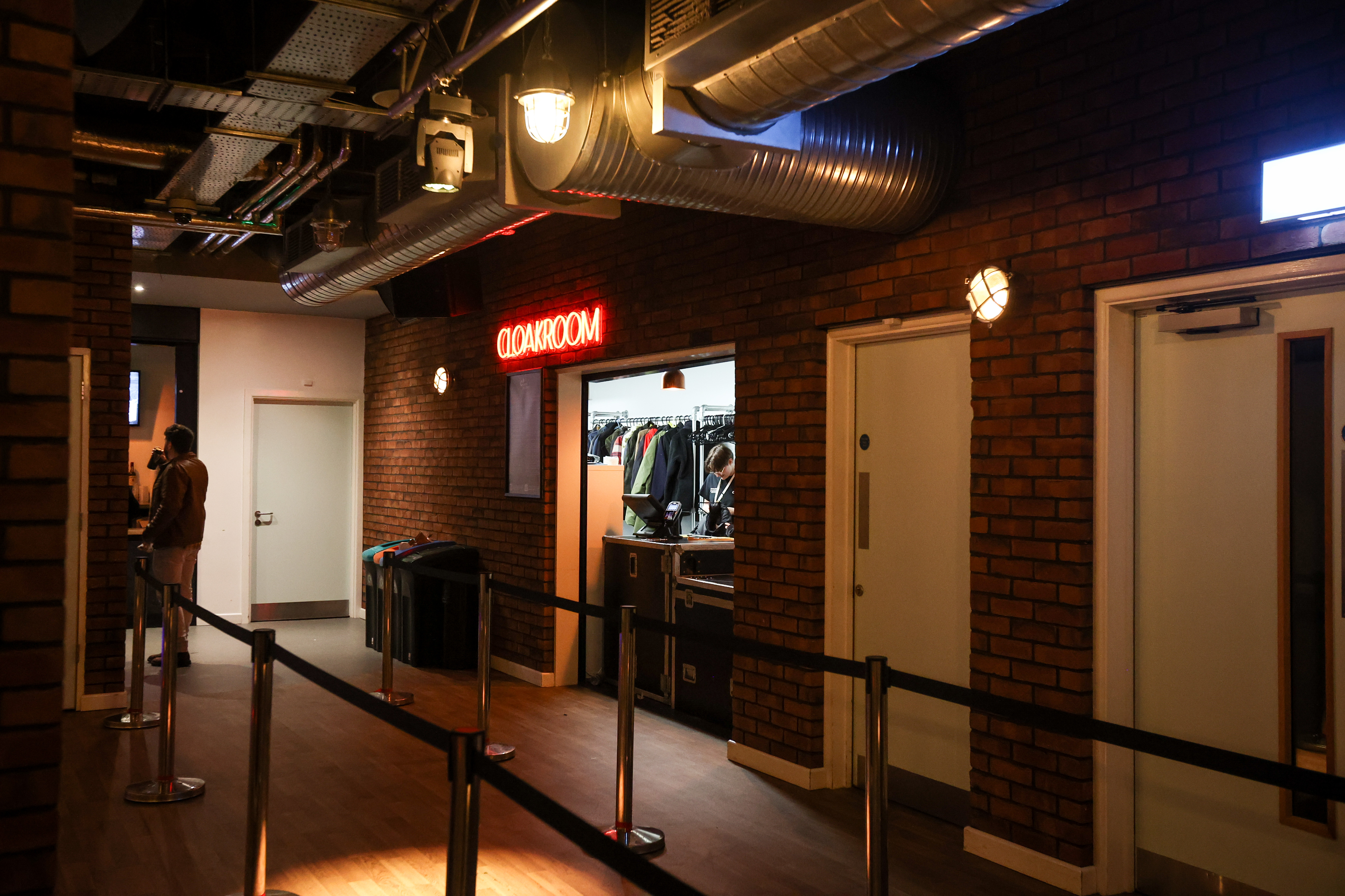A brick-walled corridor with a red neon “Cloakroom” sign above an open doorway. Inside, a staff member is sorting coats hung on racks. Black belt barriers line the way to the cloakroom, and overhead metal ducting runs along the ceiling. A person stands in the background on the left, near a bar area, and two white doors are visible to the right.