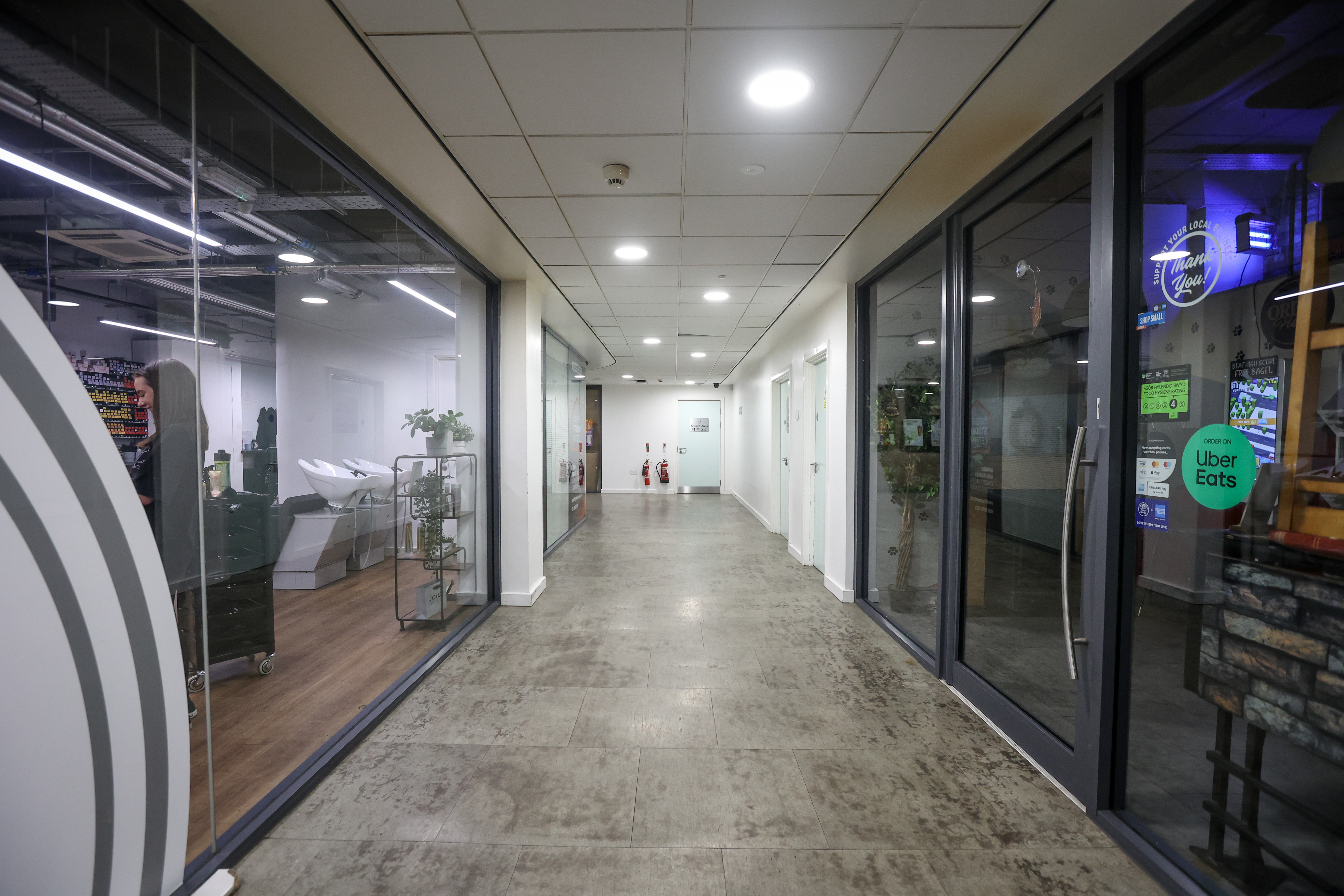 A photograph of a long, well-lit hallway showing the entrance to the venue. The hallway has glass-walled offices on both sides. The floor is a light-coloured, smooth concrete or tile. The hallway is illuminated by evenly spaced ceiling lights. A fire alarm or sensor is visible on the ceiling. The perspective of the hallway leads the viewer's eye towards the far end.