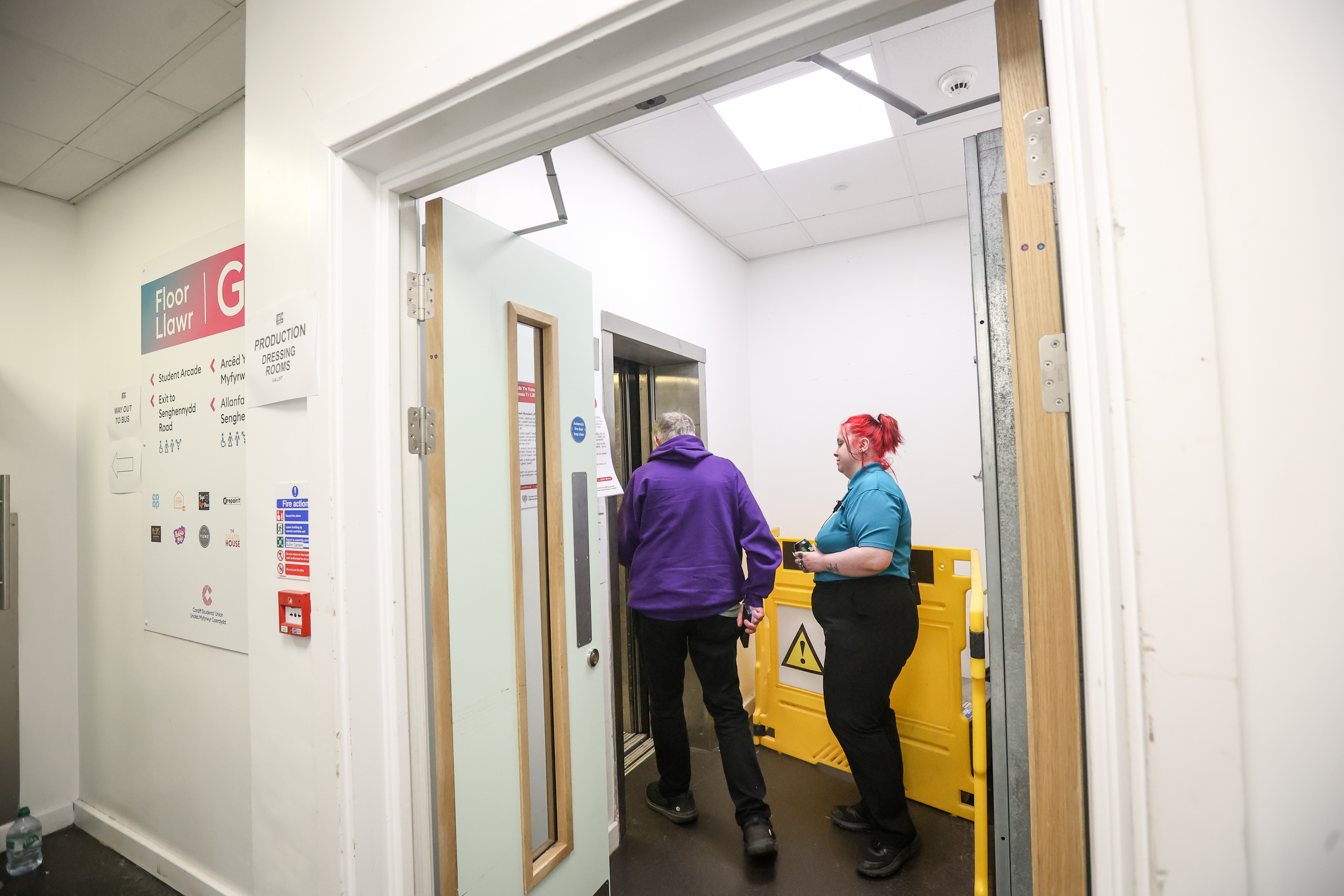 A photo showing a customer being directed to the lift. A bright, white-walled corridor on Floor G, with signage indicating directions. On the left, a large sign and fire safety notices are visible. An open lift is straight ahead, where a person in a purple hoodie step inside, accompanied by a staff member in a teal polo shirt.
