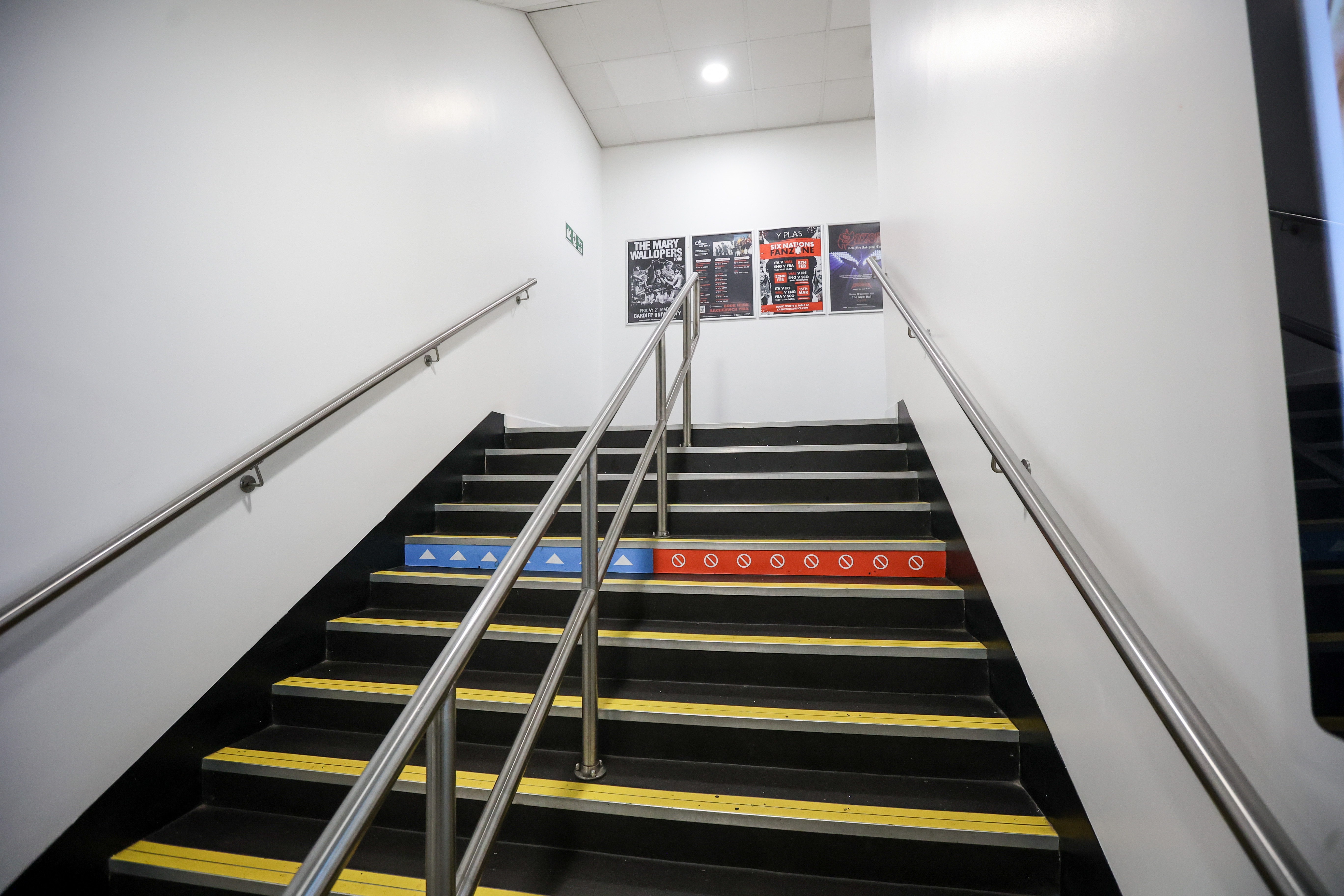 A photograph of a staircase leading upwards. The steps are black with yellow and blue striped edges, providing visual contrast. Two metal handrails run parallel to the staircase on either side. Three posters are affixed to the white wall at the top of the stairs. The staircase appears to be well-lit, with a light fixture visible on the ceiling. The wall is a plain white, and the steps appear to be in good condition.