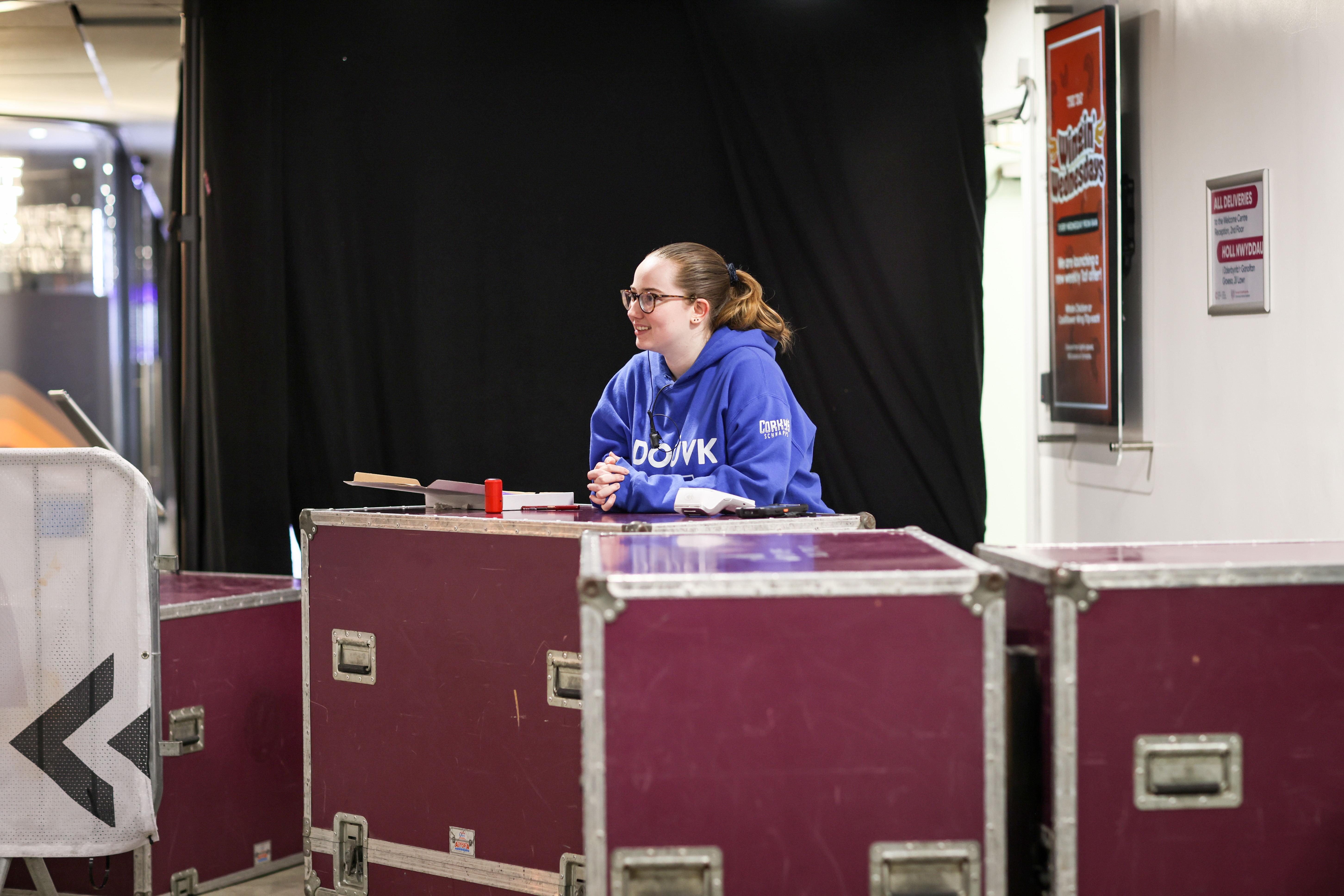A smiling staff member at the box office in a bright blue hoodie, standing behind a row of large purple flight cases used as a makeshift desk. They have their hair tied back and wears glasses. A black curtain hangs behind her, and to the right, a wall-mounted poster and sign provide event information.