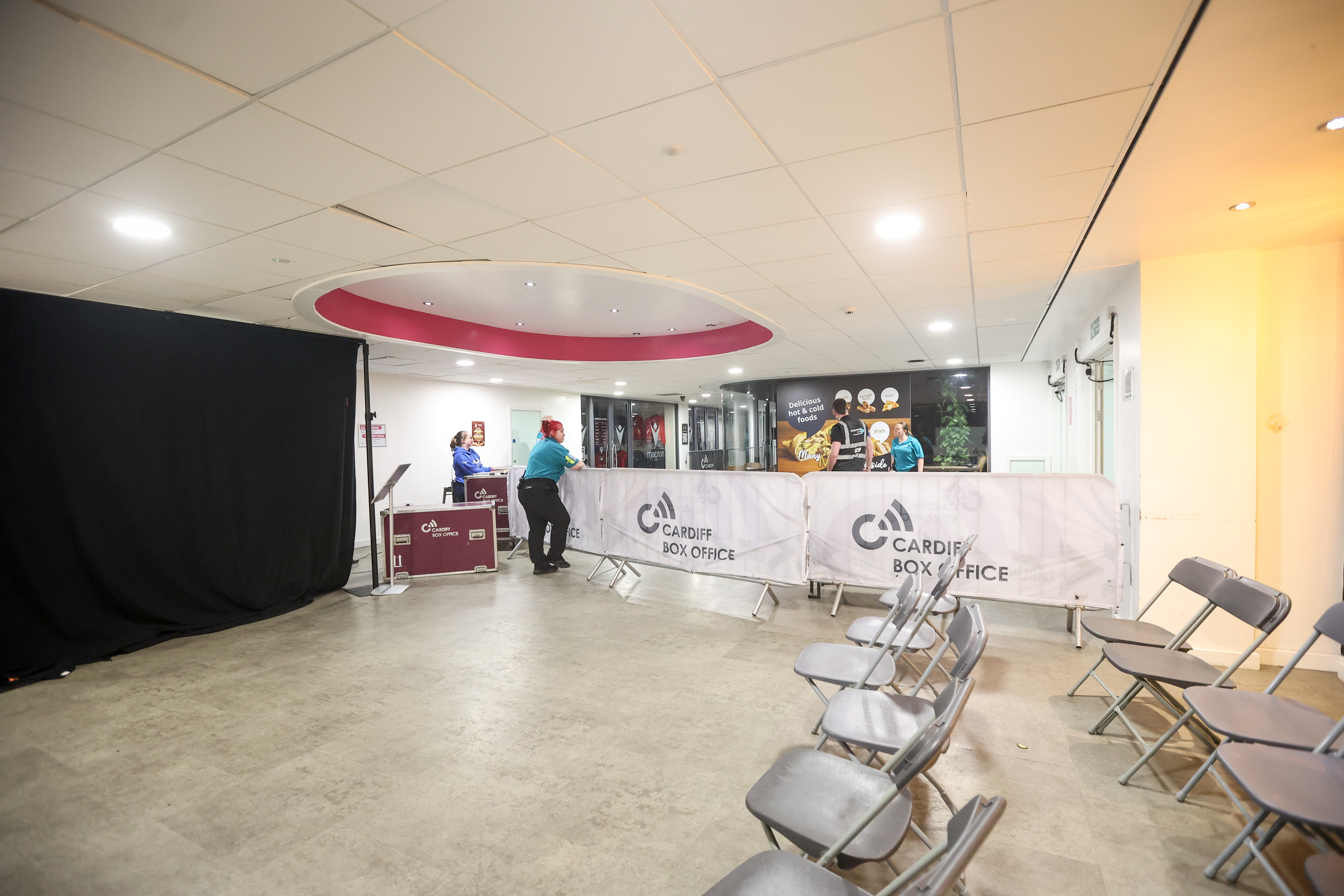 A bright lobby with a pink circular ceiling feature near the center. On the left, a large black curtain and maroon flight cases labelled “Cardiff Box Office” serve as a makeshift counter where staff in teal and blue uniforms assist customers. A white mesh barrier printed with the venue’s logo runs between the box office area and the open foyer. To the right, several grey folding chairs are set up for waiting. In the background, glass walls display posters and signage for hot and cold refreshments.