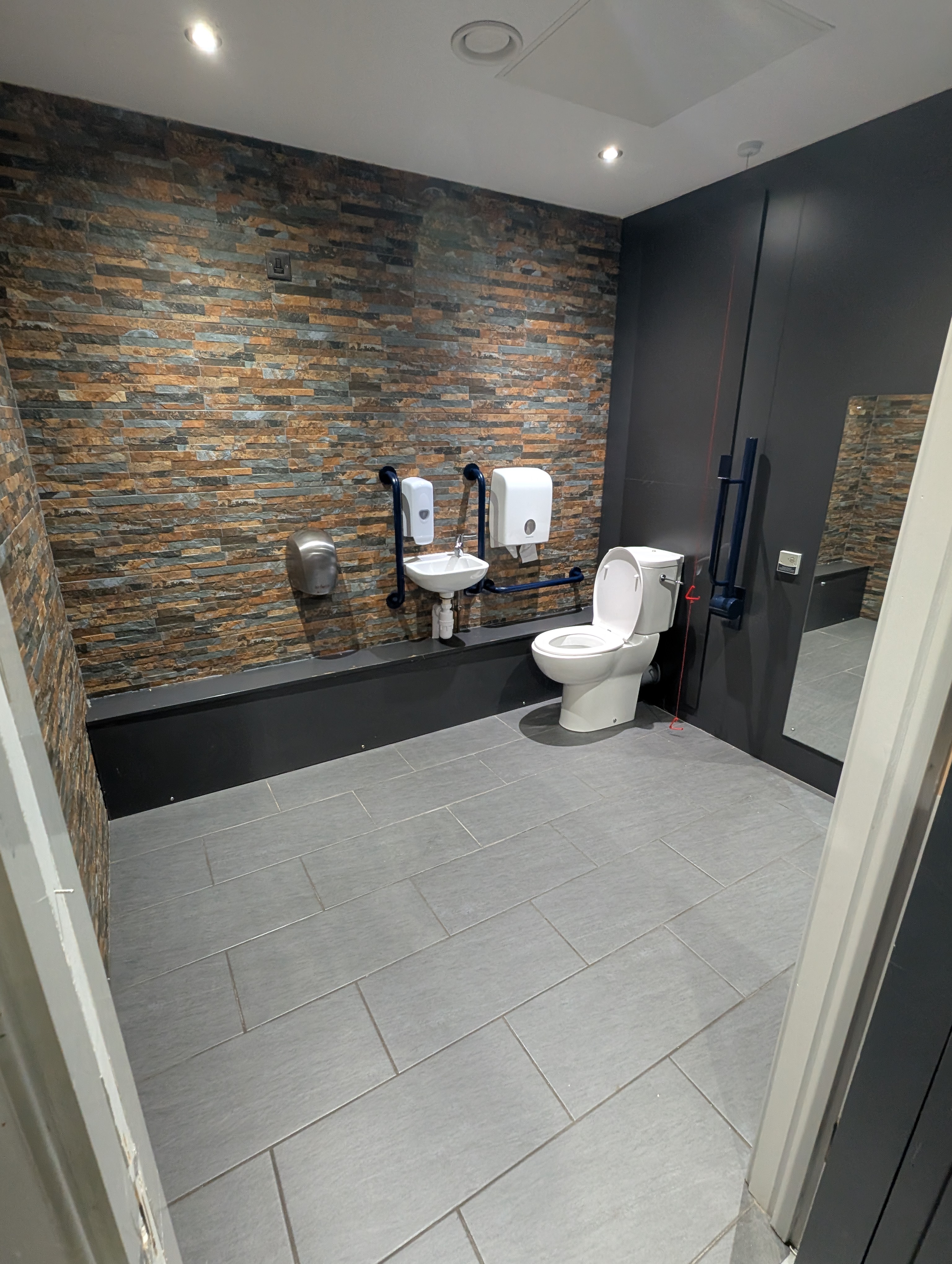 A wide, well-lit view of a modern restroom. The restroom features a white toilet, a small white sink with two vertical grab bars on either side, and a wall-mounted hand dryer. A textured accent wall behind the sink and toilet is made of multi-coloured stone-like tiles. The floor is tiled with large, rectangular grey tiles. A mirror is visible on the right wall. The restroom appears clean and well-maintained.